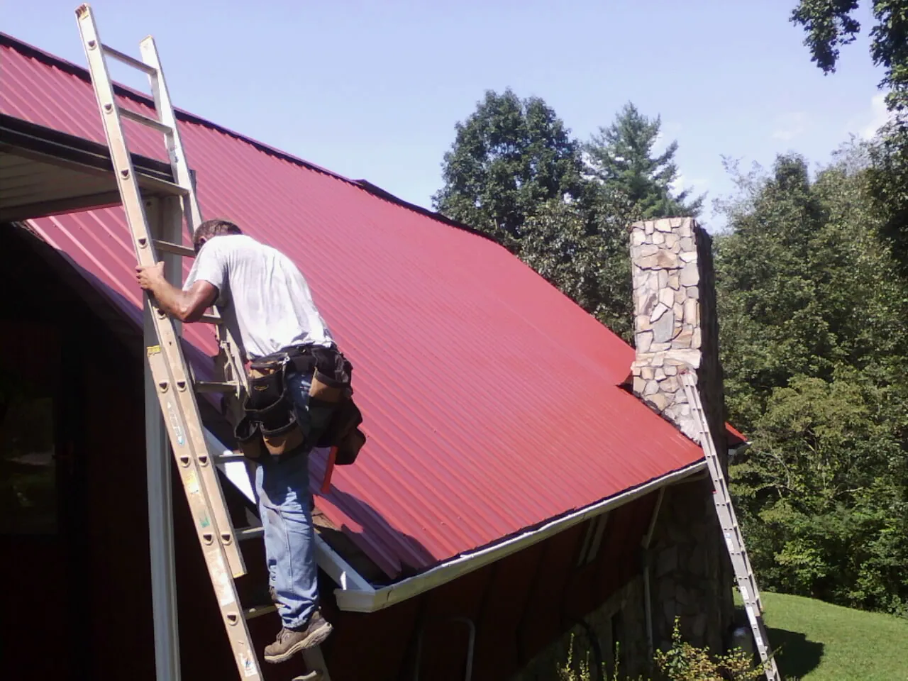 Roofer on a ladder beside a red metal roof and stone chimney — Soffit & Fascia Repair by Roofing Brothers in Tuckahoe